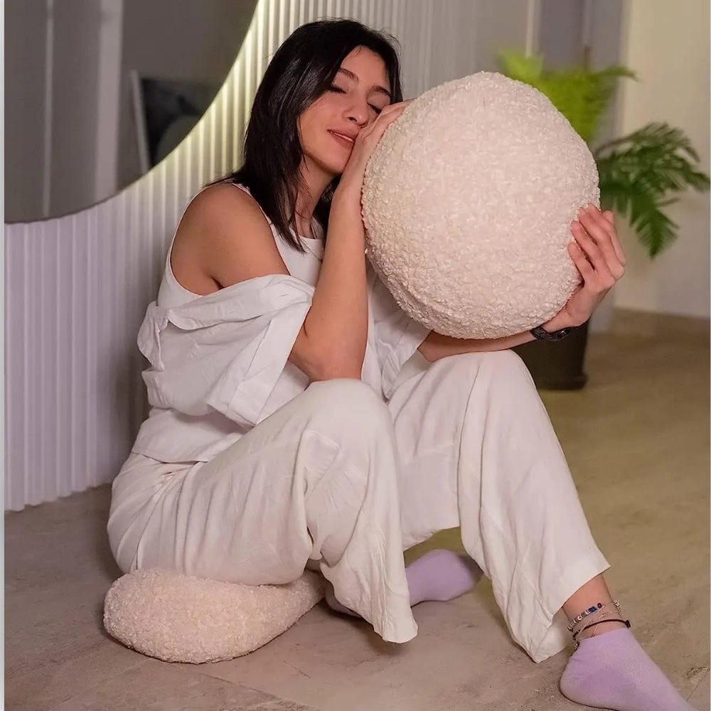 Coussin boule en matière douce et texturée, posé sur un sol en bois, avec une personne assise à côté