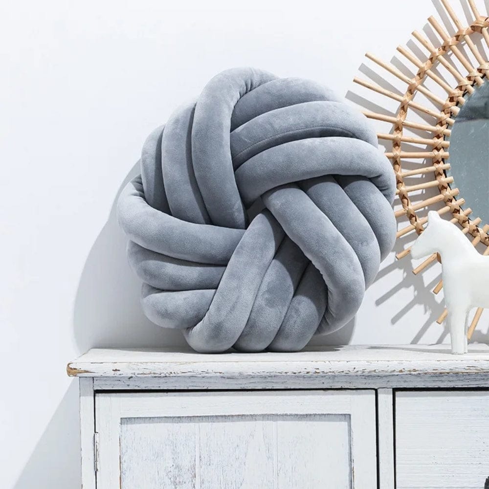 Coussin déco en velours gris en forme de nœud, posé sur un meuble blanc avec un miroir rond