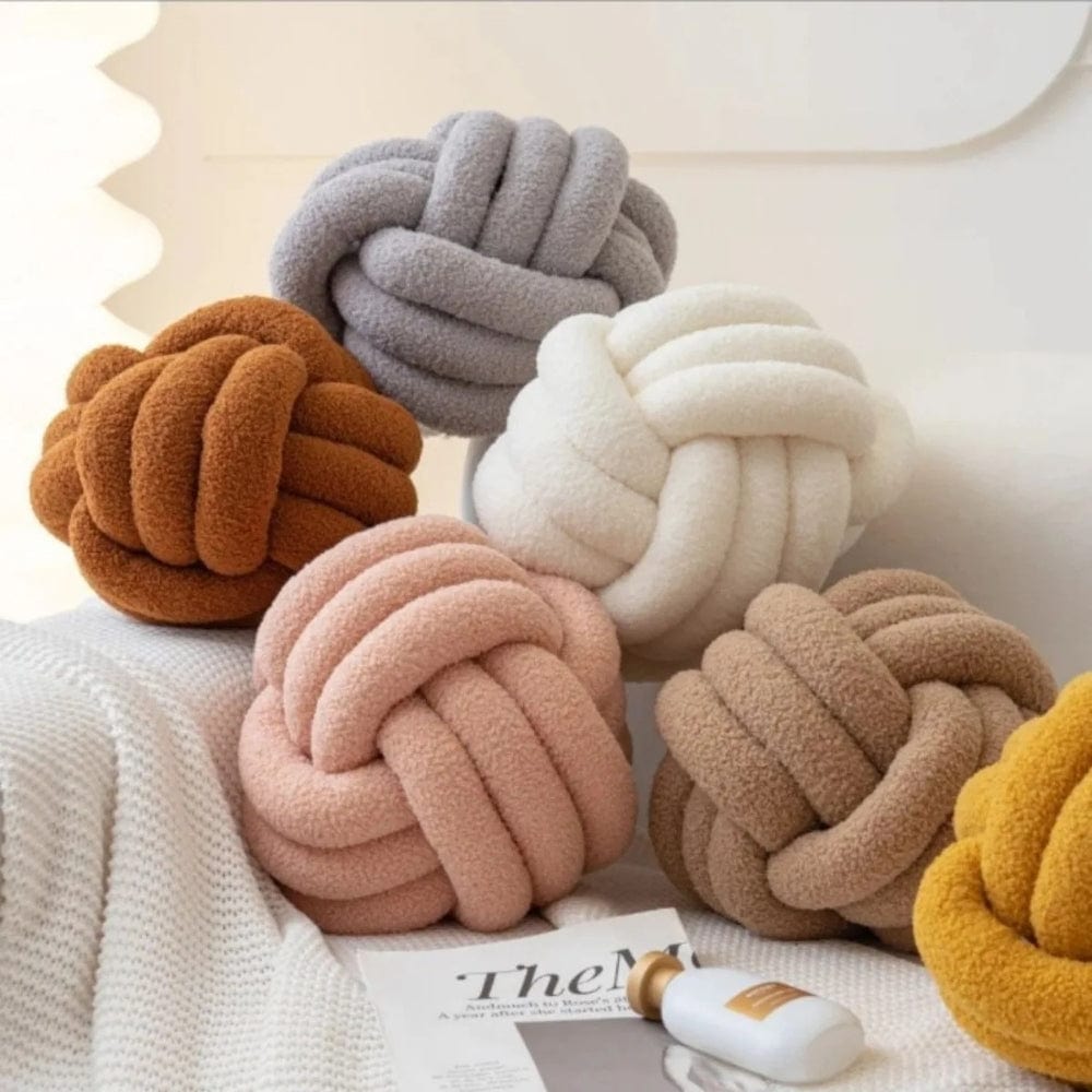 Coussins décoratifs en forme de nœud, aux teintes douces, disposés sur un canapé avec un plaid et un magazine