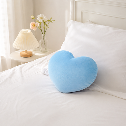 Coussin cœur bleu moelleux posé seul sur un lit blanc dans une chambre lumineuse.