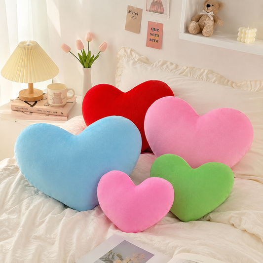 Ensemble de coussins en forme de cœur bleu, rouge, rose et vert disposés sur un lit dans une chambre lumineuse.