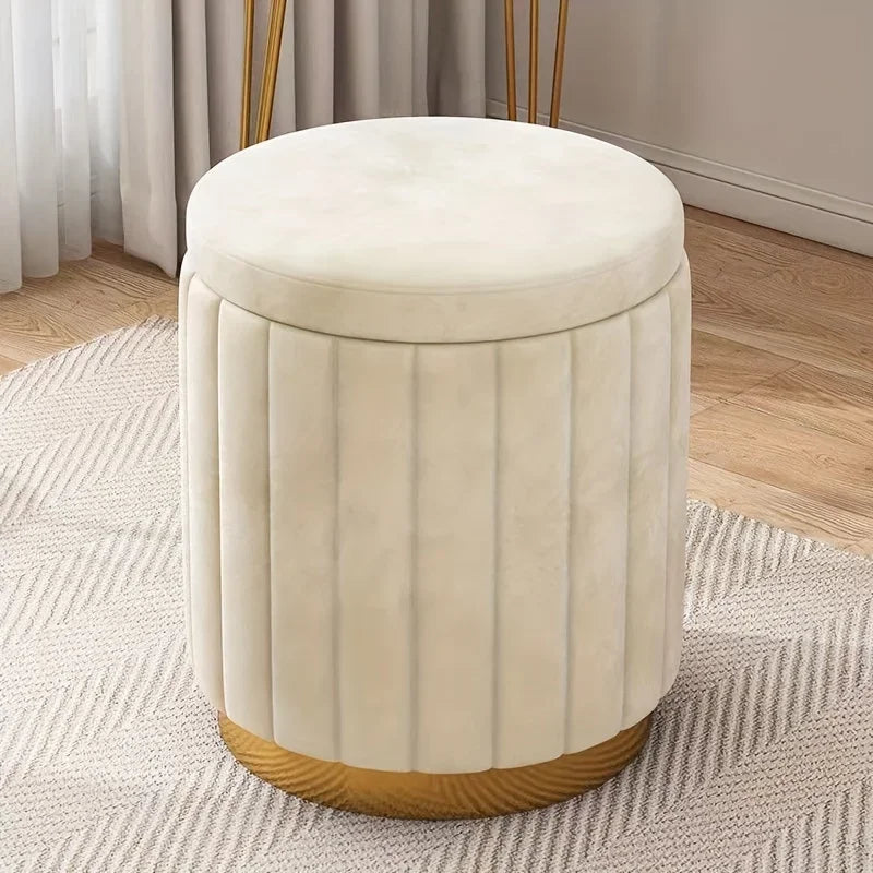 Pouf rond blanc crème en velours avec assise rembourrée et base dorée, posé sur un tapis près d’une fenêtre.