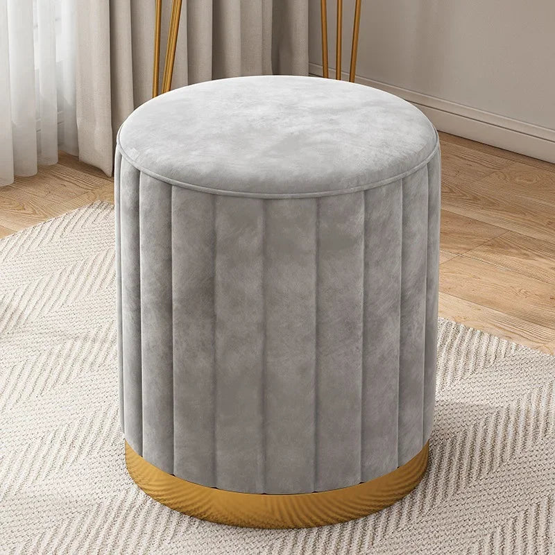 Pouf rond en velours gris avec coutures verticales et base dorée, photographié dans une pièce aux tons neutres.