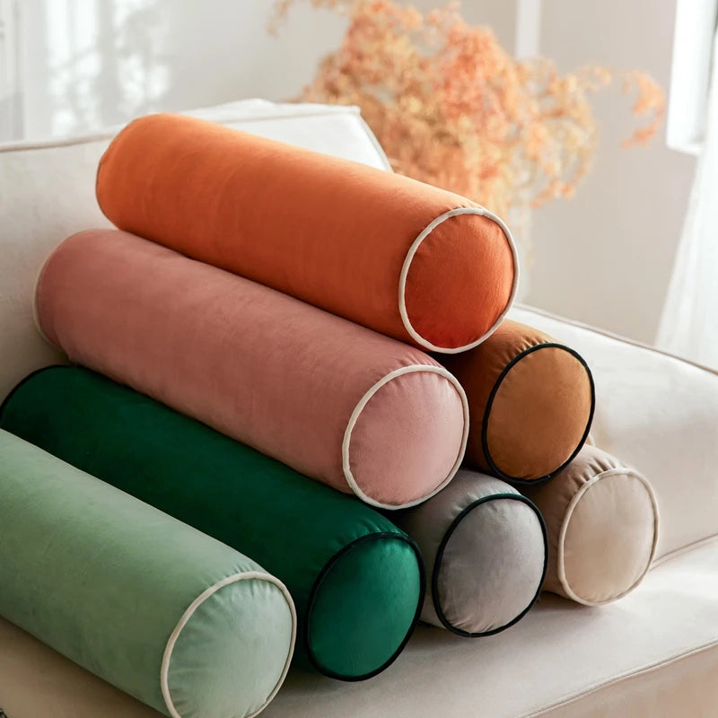 Ensemble de coussins cylindriques en velours de différentes couleurs avec passepoil contrastant, empilés sur un canapé crème.