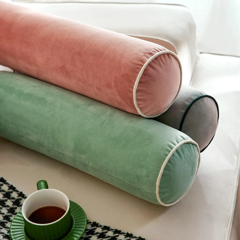 Coussins cylindriques en velours rose, vert sauge et gris posés sur un canapé près d’une tasse.
