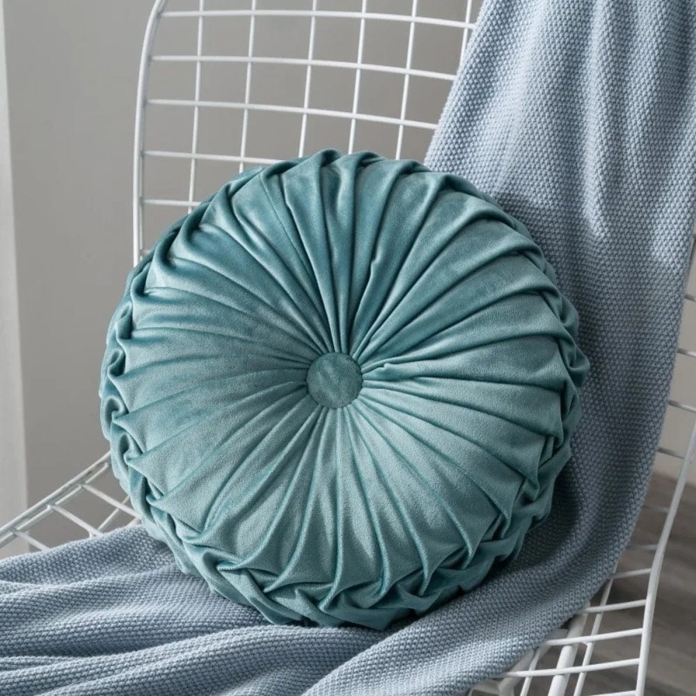 Coussin décoratif rond en velours bleu, avec des plis élégants, posé sur une chaise en métal et un plaid
