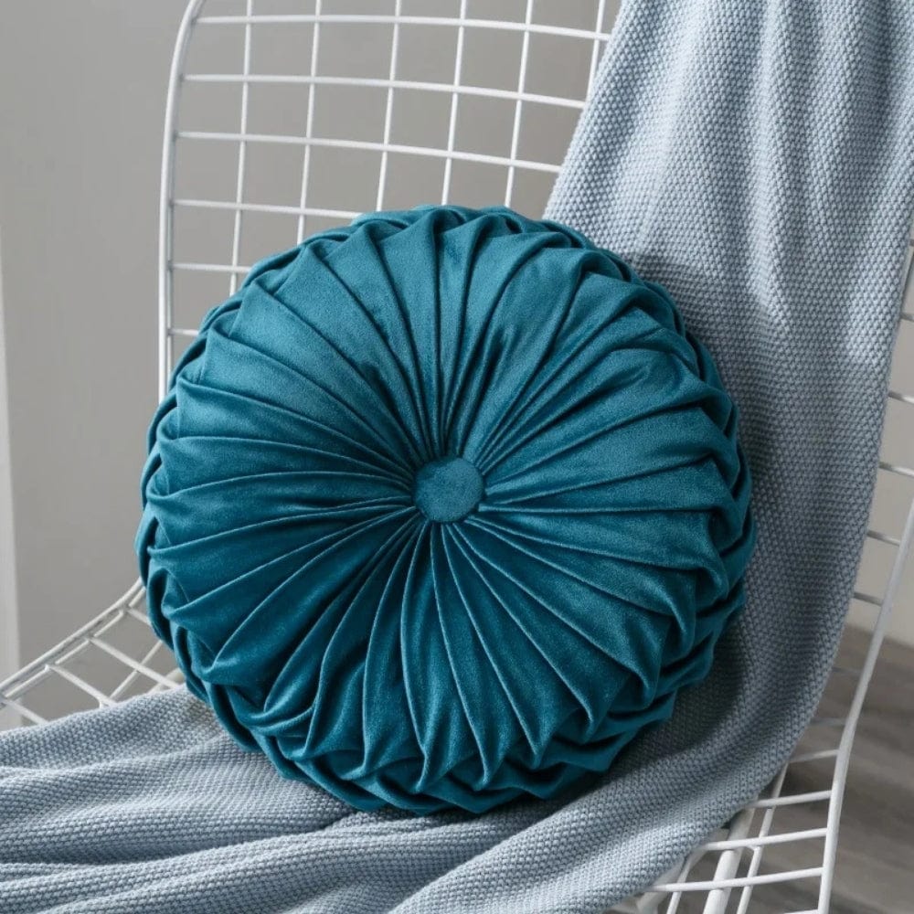 Coussin déco rond en velours bleu canard, posé sur une chaise blanche avec un plaid gris