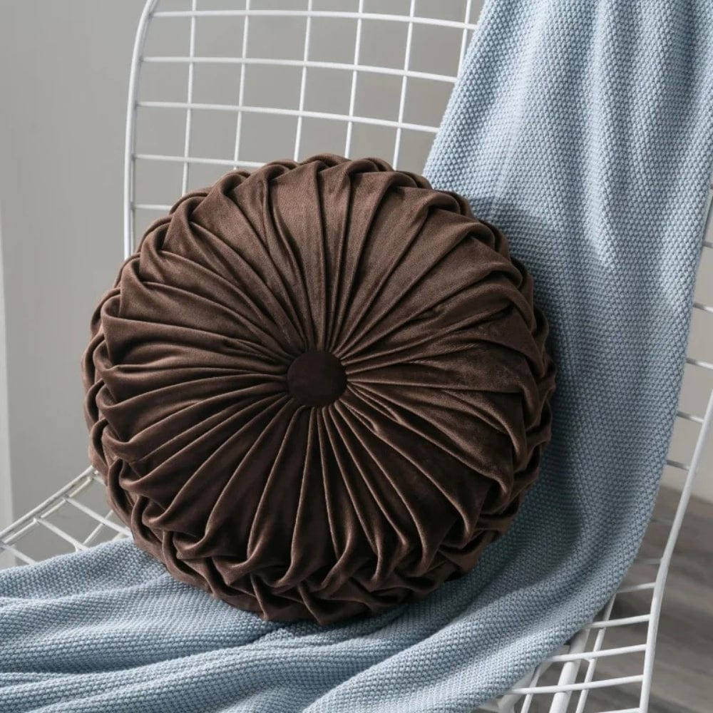 Coussin décoratif rond en velours marron, avec un motif plissé, posé sur une chaise blanche et un plaid bleu