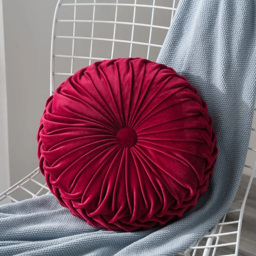 Coussin déco rond en velours rouge avec des plis, posé sur une chaise en métal, à côté d'un plaid gris