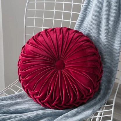 Coussin déco rond en velours rouge avec des plis, posé sur une chaise en métal, à côté d'un plaid gris