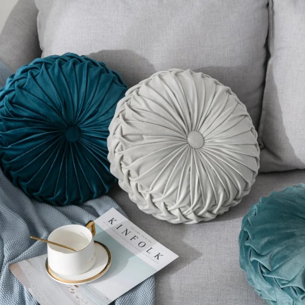 Coussins décoratifs ronds en velours gris et bleu, posés sur un canapé gris avec une tasse et un magazine
