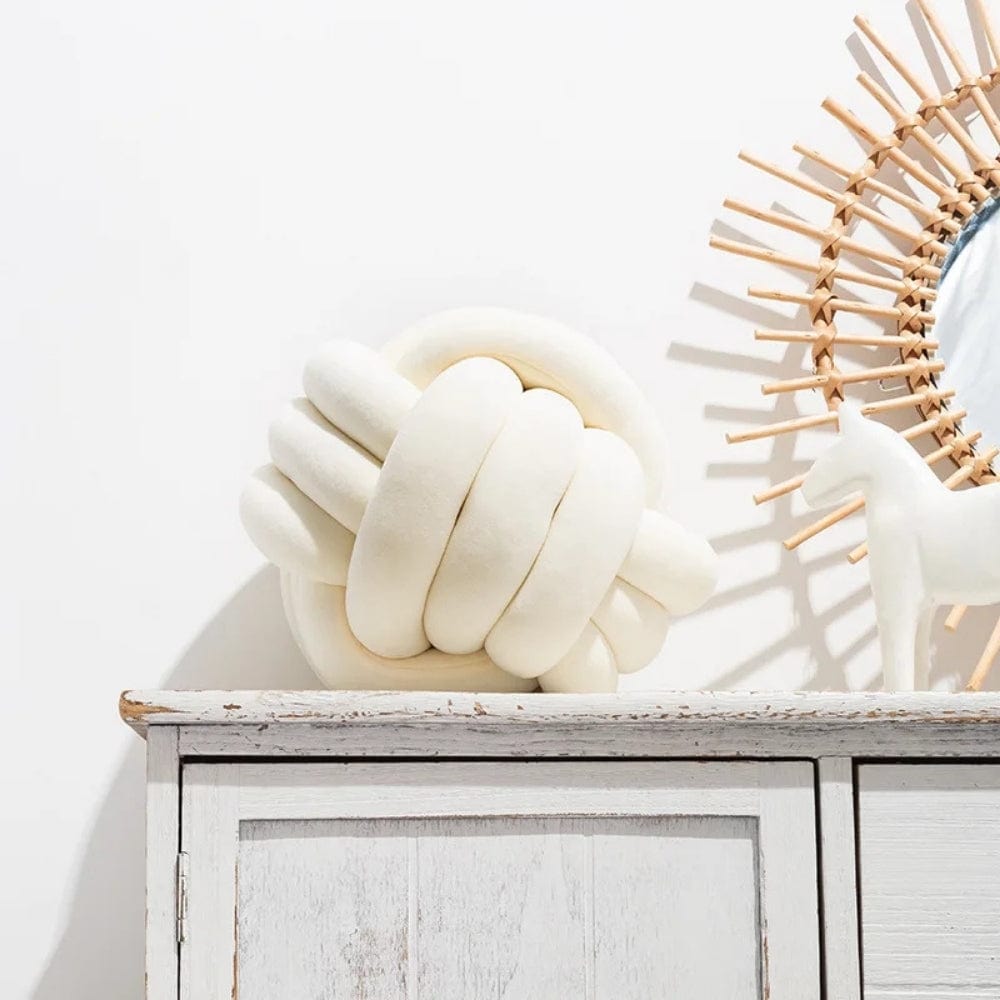 Coussin décoratif en forme de nœud, couleur blanche, posé sur un meuble en bois avec un miroir rond