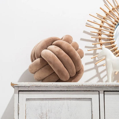 Coussin décoratif en forme de nœud, couleur beige, posé sur un meuble en bois blanc, avec un miroir en arrière-plan
