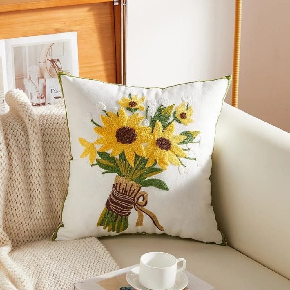 Coussin déco blanc brodé avec des tournesols sur un canapé, accompagné d'un plaid et d'une tasse
