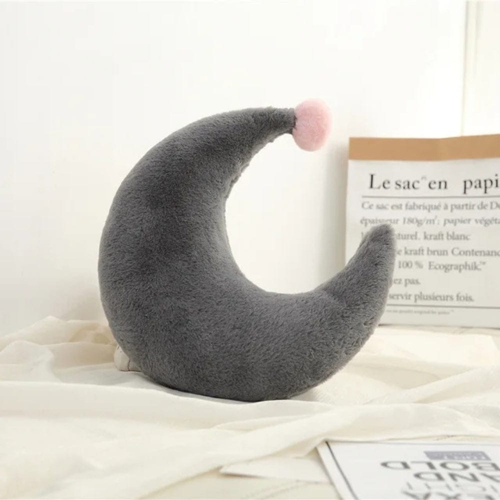 Coussin décoratif en forme de lune gris avec un pompon rose, posé sur un tissu beige