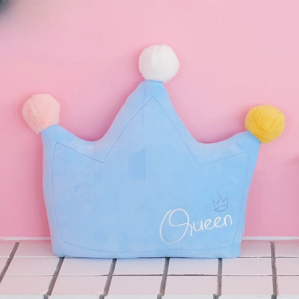 Coussin décoratif en forme de couronne, bleu avec pompons pastel, posé sur un fond rose