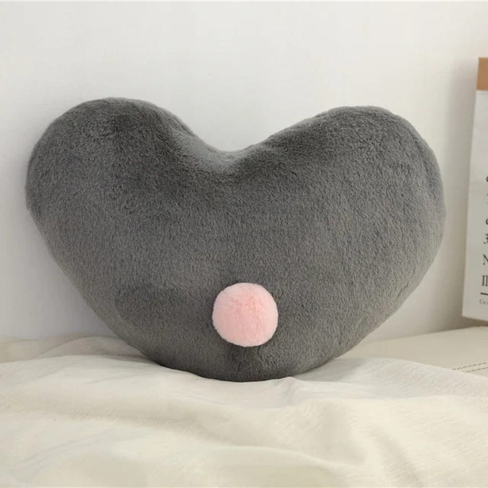 Coussin décoratif en forme de cœur gris avec un pompon rose, posé sur un lit aux draps clairs