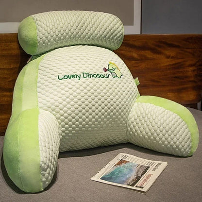 Coussin de lecture en forme de dinosaure, vert clair et blanc, posé sur un lit avec un magazine à côté