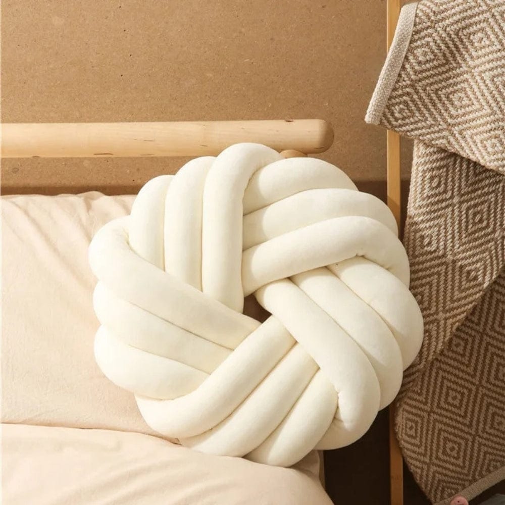 Coussin de sol en forme de nœud, couleur crème, posé sur un lit avec une couverture en motif géométrique