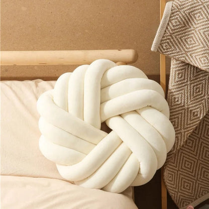 Coussin de sol en forme de nœud, couleur crème, posé sur un lit avec une couverture en motif géométrique