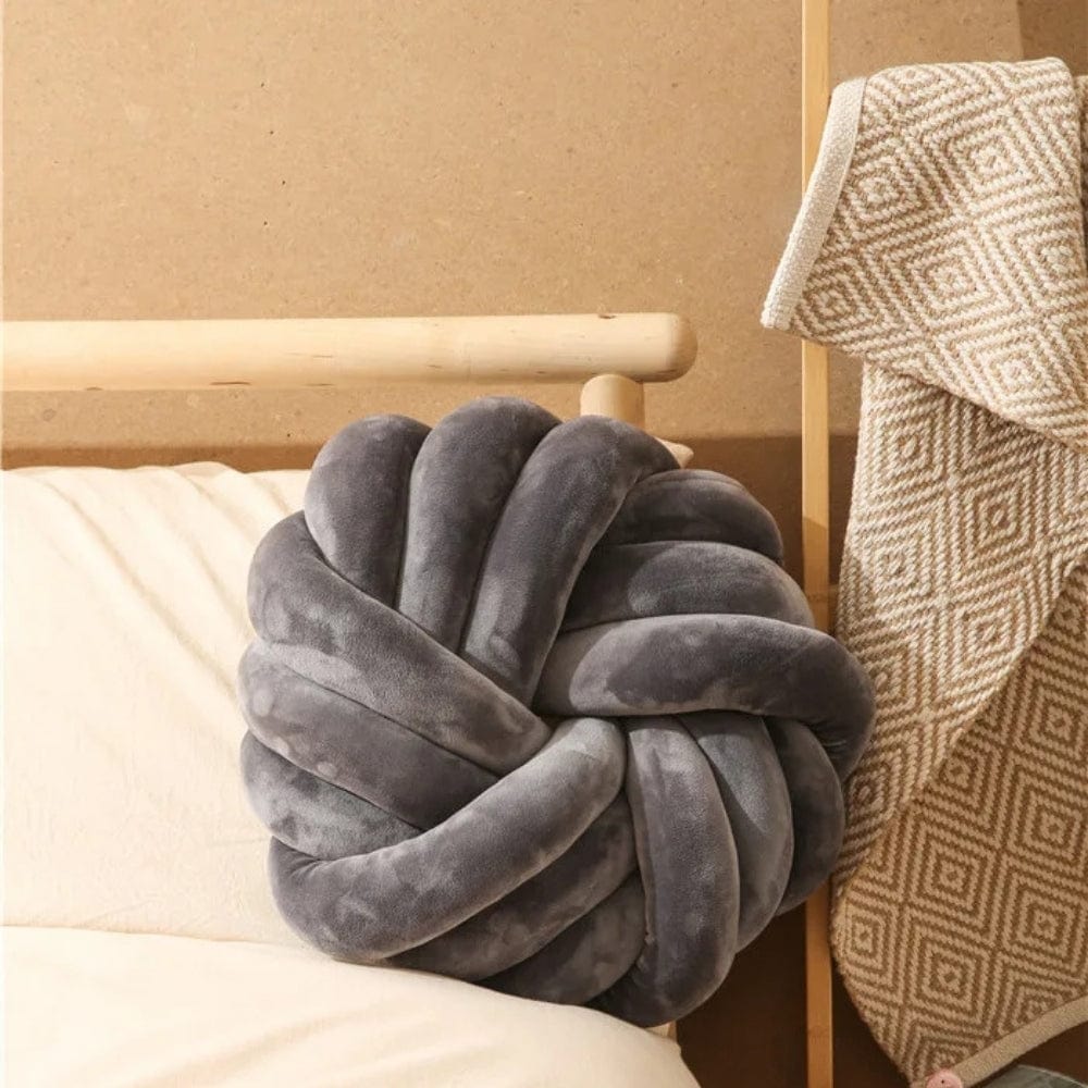 Coussin de sol en velours gris en forme de nœud, posé sur un lit avec un plaid décoratif