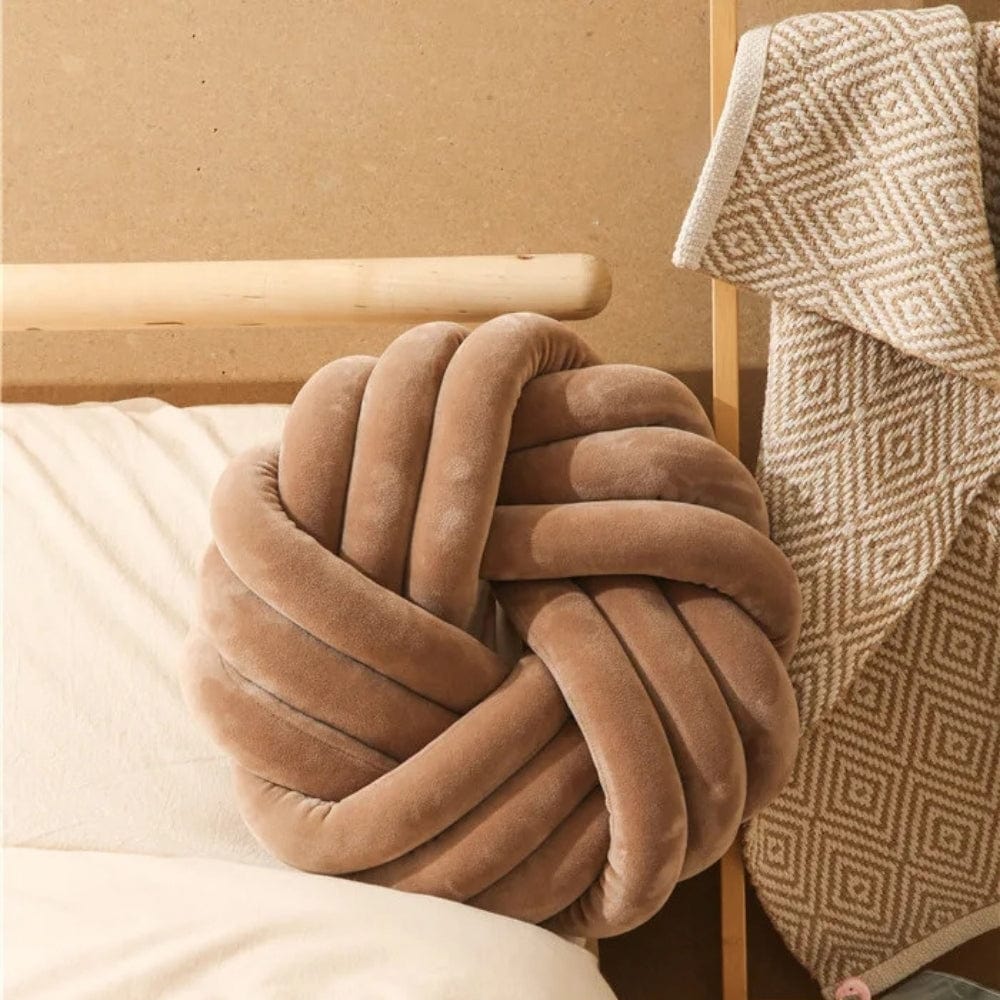 Coussin déco en velours beige en forme de nœud, posé sur un lit avec une couverture à motifs