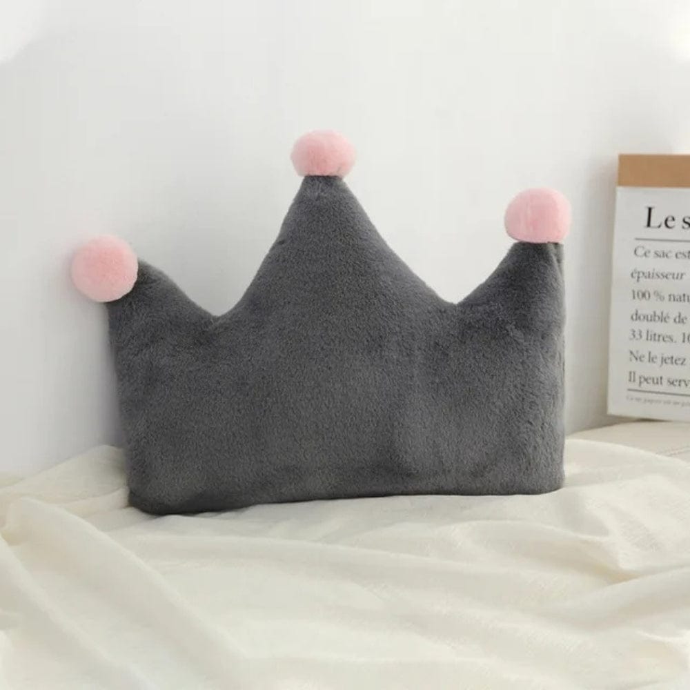 Coussin décoratif en forme de couronne gris avec pompons roses, posé sur un lit aux draps blancs