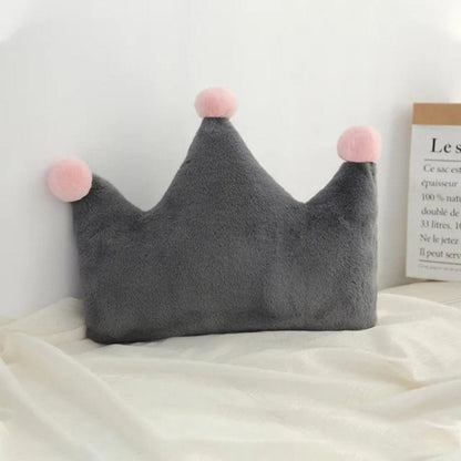 Coussin décoratif en forme de couronne gris avec pompons roses, posé sur un lit aux draps blancs