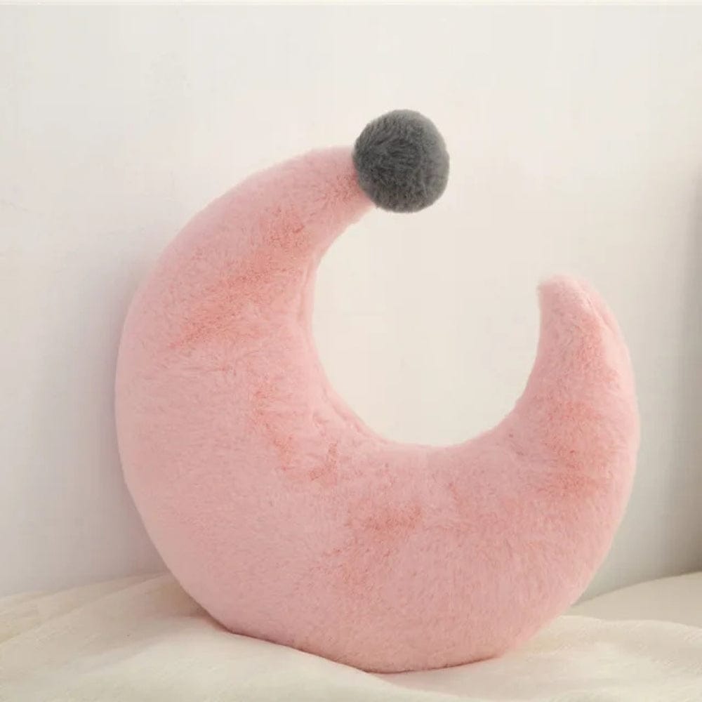 Coussin déco en forme de croissant, doux et rose, avec un pompon gris, posé sur un lit