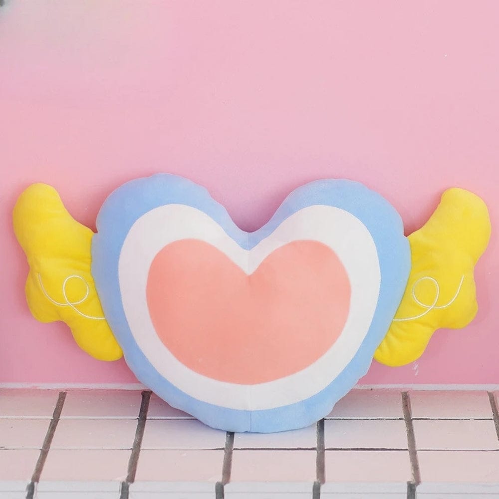 Coussin en forme de cœur aux couleurs pastel, avec des ailes jaunes, posé sur un fond rose