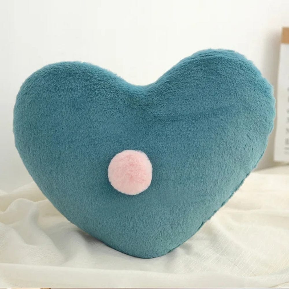 Coussin décoratif en forme de cœur, en velours bleu avec un pompon rose, posé sur un tissu neutre