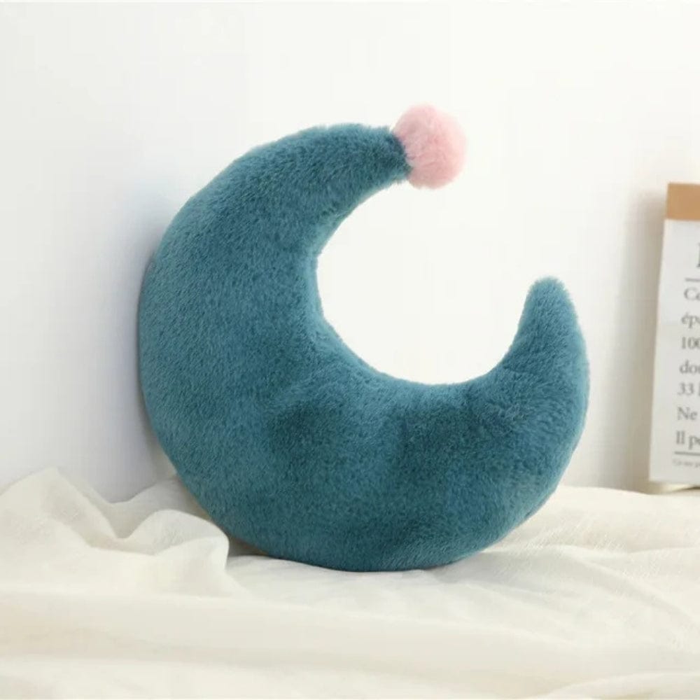 Coussin décoratif en forme de lune, en velours bleu avec un pompon rose, posé sur un tissu clair