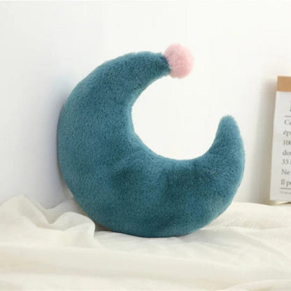 Coussin décoratif en forme de lune, en velours bleu avec un pompon rose, posé sur un tissu clair