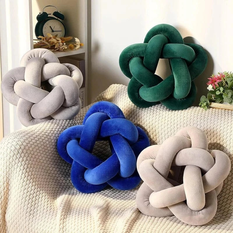 Quatre coussins décoratifs en velours, de formes nouées, posés sur un plaid dans un salon lumineux