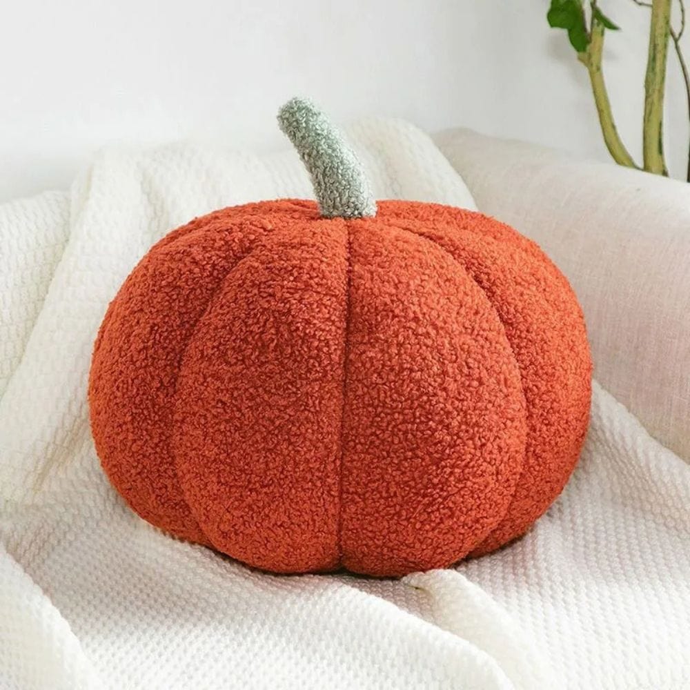 Coussin décoratif en forme de citrouille orange avec une tige verte, posé sur un plaid blanc