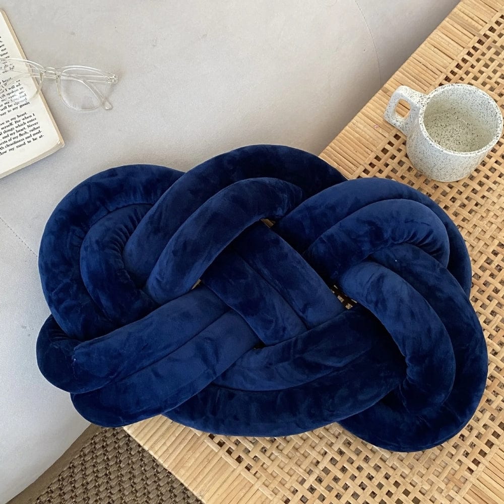 Coussin décoratif en velours bleu nuit, en forme de nœud, posé sur une table en bois tressé