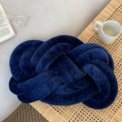 Coussin décoratif en velours bleu nuit, en forme de nœud, posé sur une table en bois tressé