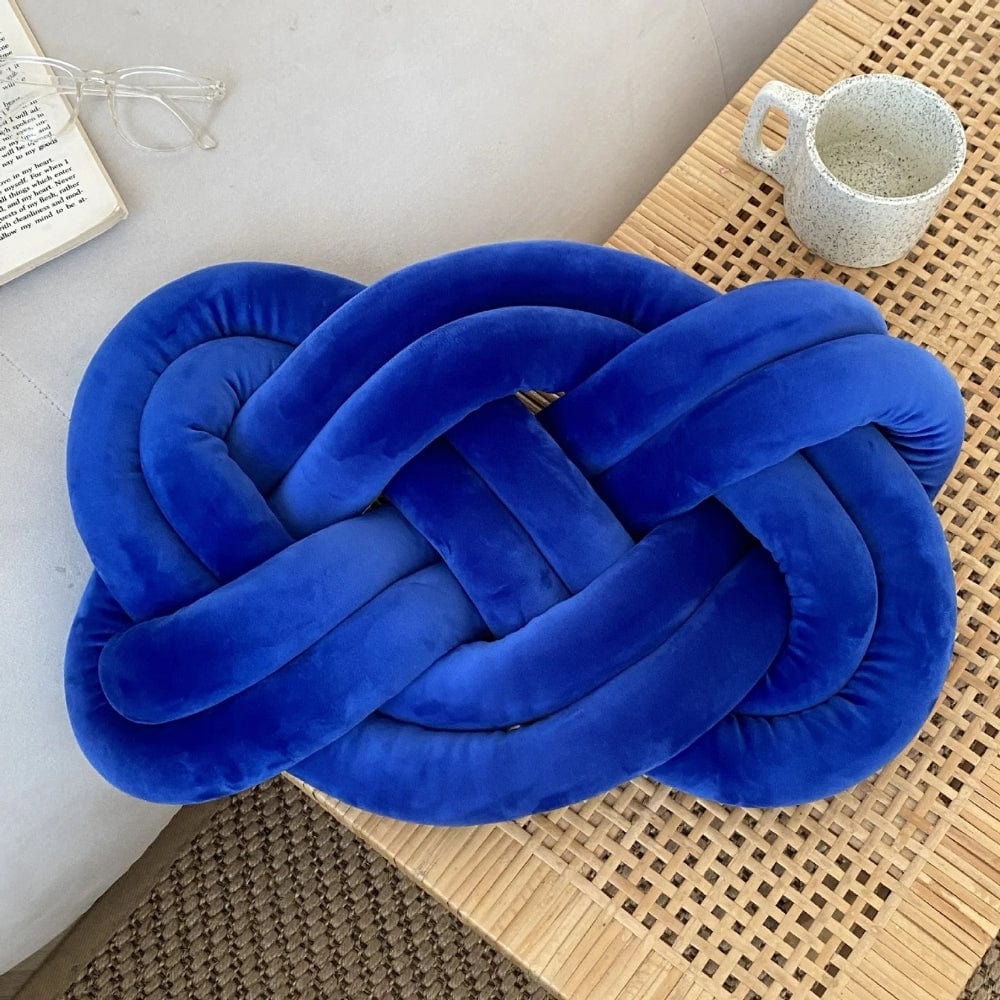 Coussin décoratif en velours bleu en forme de nœud, posé sur une table en bois, à côté d'une tasse