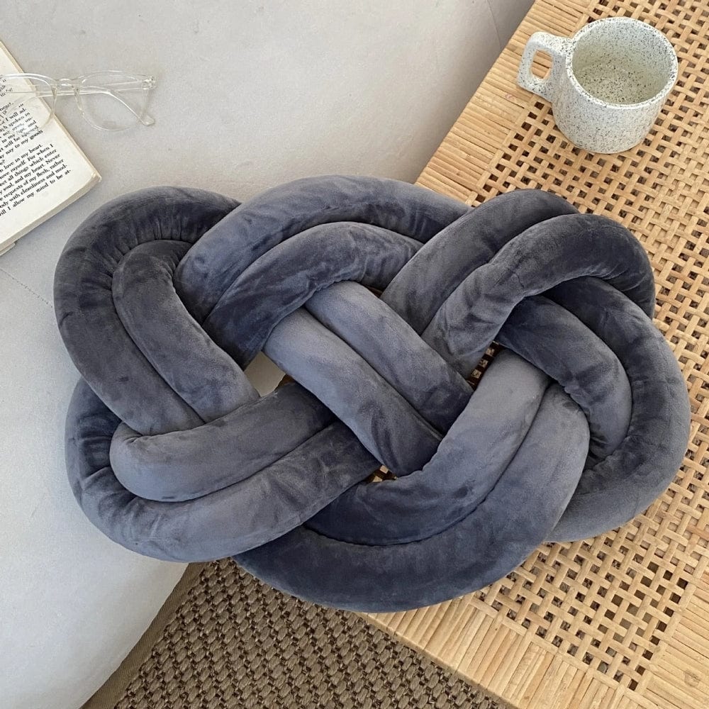 Coussin décoratif en velours gris en forme de nœud, posé sur une table en bois, apportant une touche moderne au