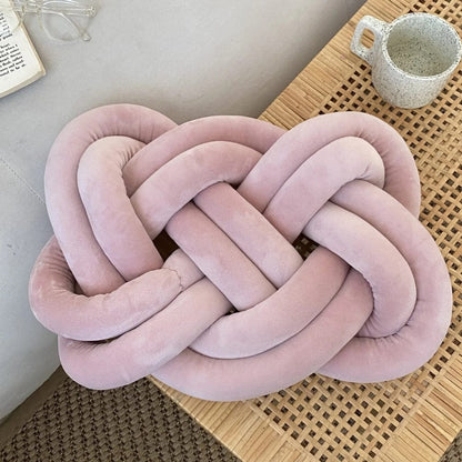 Coussin décoratif en velours rose en forme de nœud, posé sur une table en bois, avec une tasse à côté