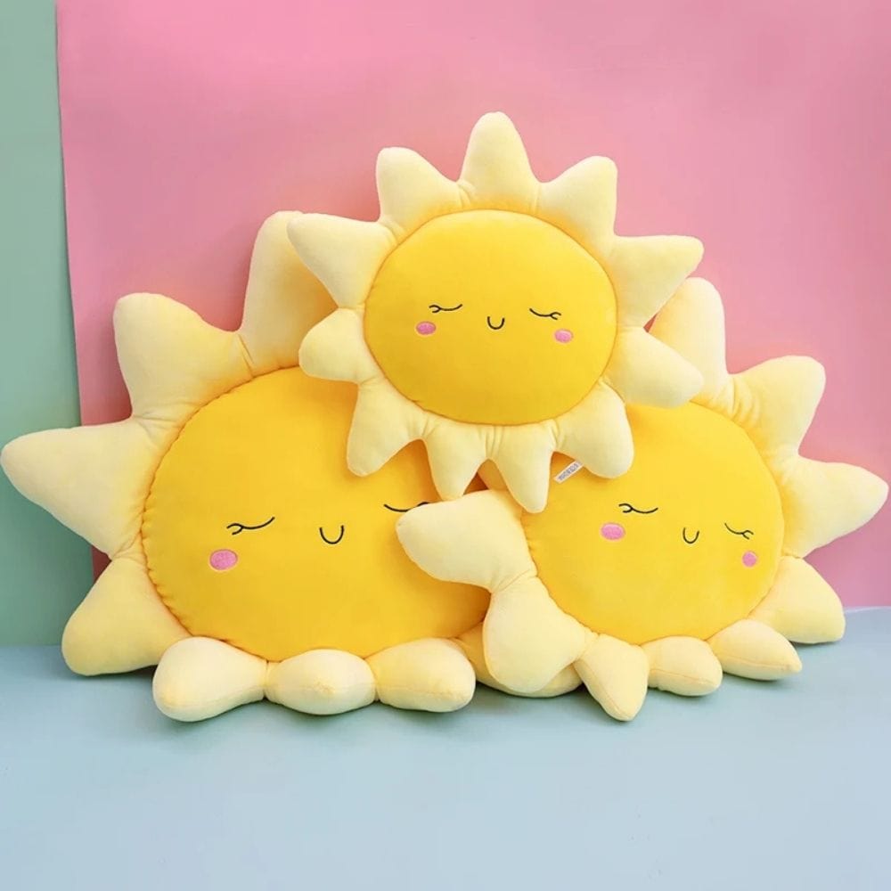 Trois coussins en peluche jaune en forme de soleil, posés sur une surface bleue, avec un fond rose