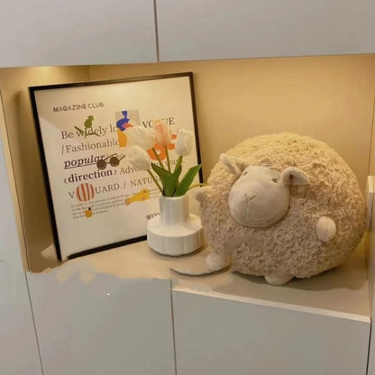 Coussin décoratif en forme de mouton, couleur beige, posé sur une étagère avec un vase et un tableau