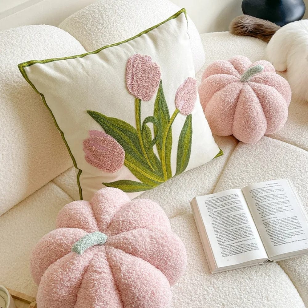 Coussin décoratif beige avec des tulipes roses, posé sur un canapé blanc, entouré de coussins en forme de citrouille