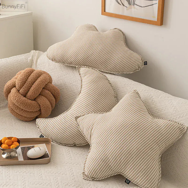 Coussins décoratifs en forme d'étoile et de lune, rayés beige sur un canapé blanc, ajoutant une touche chaleureuse à la