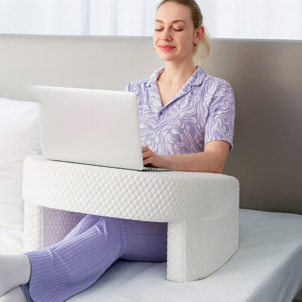 Coussin blanc en forme de support, posé sur un lit, avec une femme utilisant un ordinateur portable