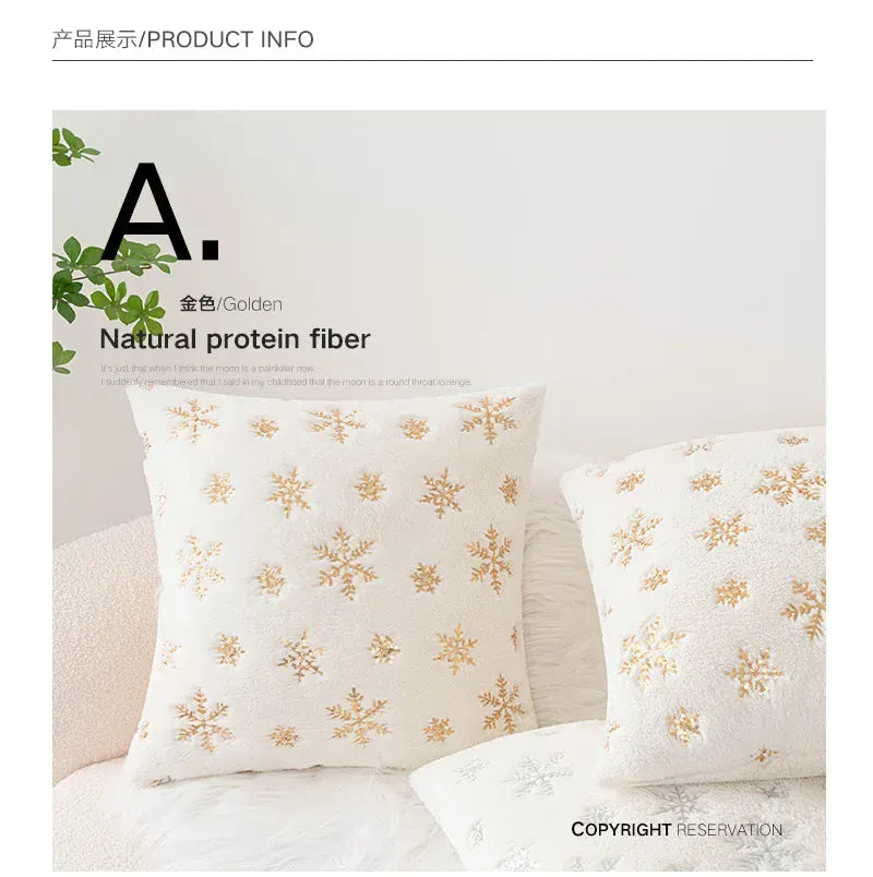Coussin décoratif blanc avec motifs dorés de flocons de neige, posé sur un canapé en tissu doux