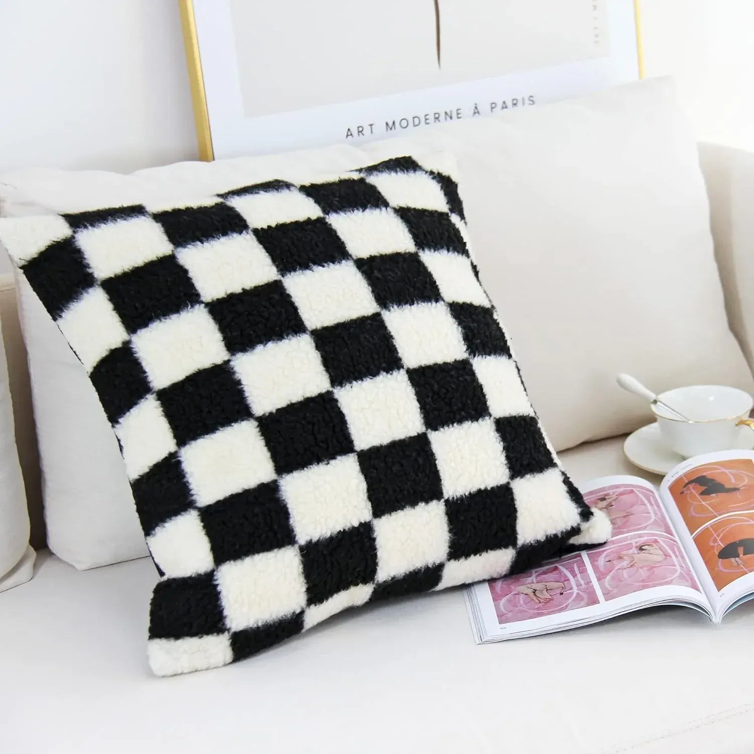 Coussin décoratif à motifs carreaux noir et blanc, posé sur un canapé, avec un livre et une tasse à côté