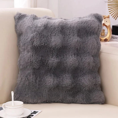 Coussin décoratif en fausse fourrure grise, posé sur un canapé beige, apportant une touche cocooning à l'intérieur
