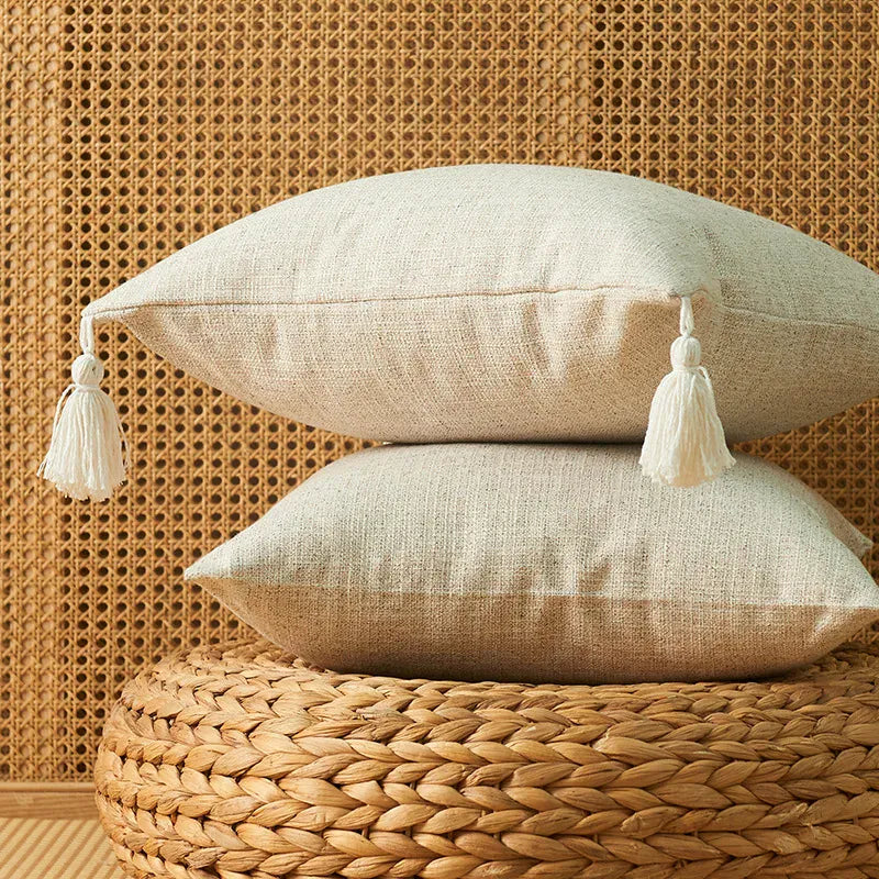 Housse de coussin beige avec pompon, posée sur un pouf en osier, décorant un intérieur chaleureux