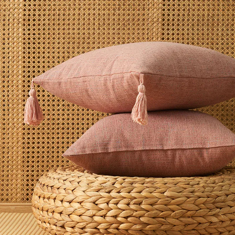 Deux coussins décoratifs rose avec pompons, posés sur un pouf en osier, devant un fond en rotin